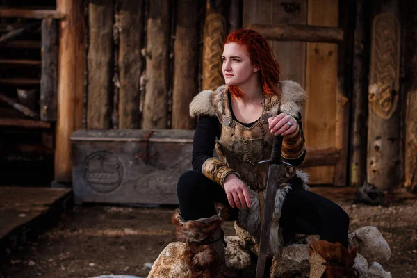 Red-haired woman is a Viking. Against the backdrop of a village or an ancient settlement. In the hands of a sword — Stock Photo, Image