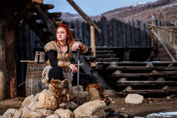 Red-haired woman is a Viking. Against the backdrop of a village or an ancient settlement. In the hands of a sword — Stock Photo, Image