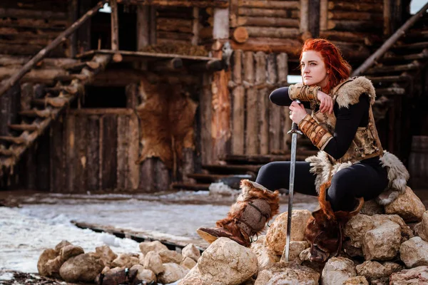 Kızıl saçlı kadın bir Viking var. Bir köy veya antik bir yerleşim fonunda. Bir kılıç elinde — Stok fotoğraf
