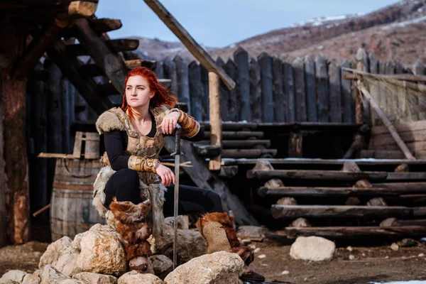 Red-haired woman is a Viking. Against the backdrop of a village or an ancient settlement. In the hands of a sword — Stock Photo, Image