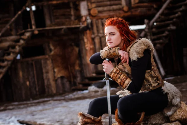 Rödhårig kvinna är en Viking. Mot bakgrund av en by eller en forntida bosättning. I händerna på ett svärd — Stockfoto