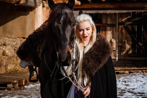 Vacker ung blond på en kråka. Kvinna viking med en svart häst mot bakgrund av berg — Stockfoto