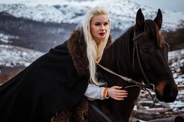 Beautiful young blonde on a crow. Woman viking with a black horse against the background of mountains — Stock Photo, Image