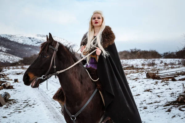 Beautiful young blonde on a crow. Woman viking with a black horse against the background of mountains — Stock Photo, Image