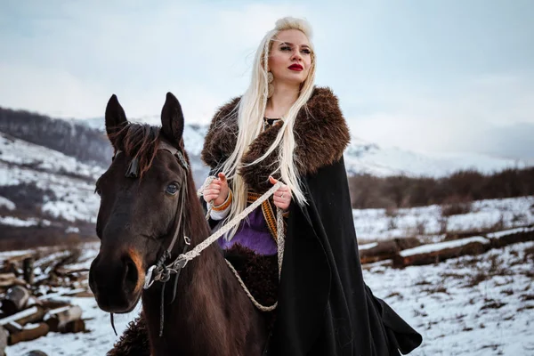 Beautiful young blonde on a crow. Woman viking with a black horse against the background of mountains — Stock Photo, Image