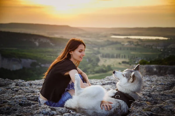 Schönes Mädchen spielt mit einem Hund, einem grauen und weißen Husky, in den Bergen bei Sonnenuntergang — Stockfoto