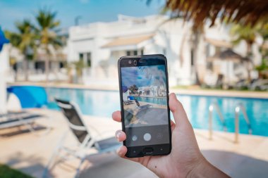 Otelde havuzun arka planda el tutarak telefon. ekranda fotoğraf kamerası.