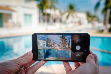 Otelde havuzun arka planda el tutarak telefon. ekranda fotoğraf kamerası.
