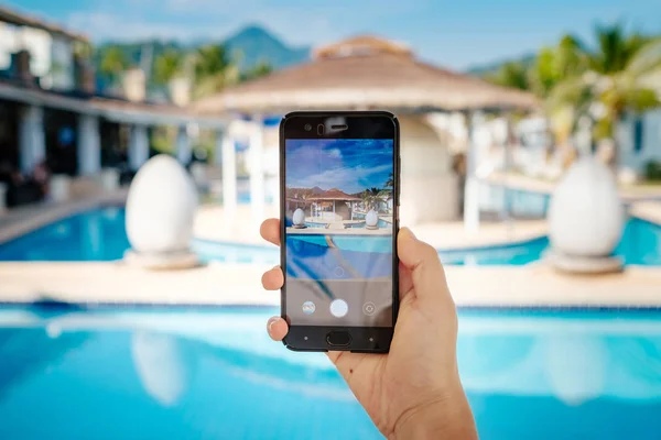 Ręcznie trzymając telefon na tle basenu w hotelu. aparat fotograficzny na ekranie. — Zdjęcie stockowe