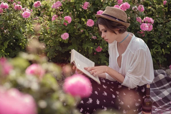 Красива дівчина в капелюсі з книгою, що сидить на траві в рожевому гадені — стокове фото