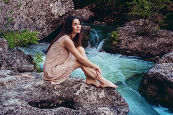 Jovem mulher no vestido está sentado na pedra no meio de um riacho . — Fotografia de Stock