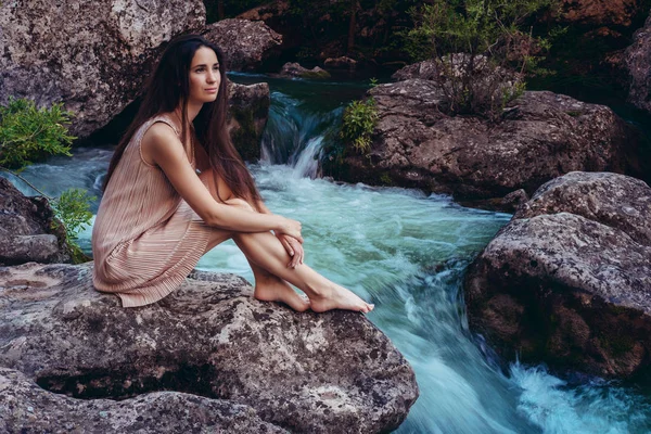 Jovem mulher no vestido está sentado na pedra no meio de um riacho . — Fotografia de Stock