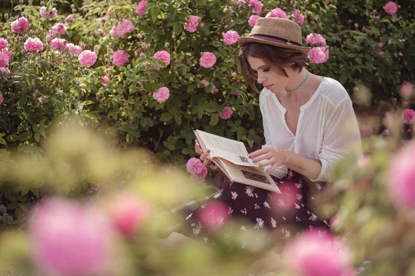 Hermosa chica usando sombrero con libro sentado en la hierba en rosa gaden —  Fotos de Stock
