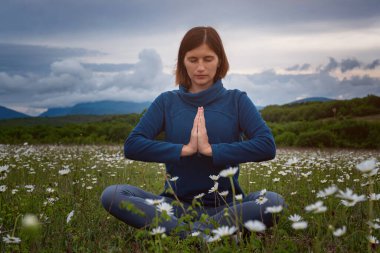 Sahada yoga yapan genç bir kadın. Gün batımından önce bir papatya tarlasında meditasyon. Koyu mavi renkler, sakin ve huzurlu. Ruhani ve duygusal kavram.