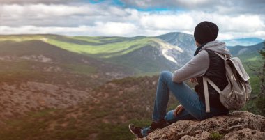 Şapka takan ve dağ uçurumunun tepesinde oturan genç bir kadın rahatlatıcı bir ruh haliyle ormanın, mavi gökyüzünün ve tatildeki bulutların güzel manzarasını seyrediyor. Asyalı kadın yalnız seyahat eder..