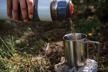 Kamp bardağında dişi bir el, doğadaki bir termostan çay doldurur. Mola, dinlenme ve seyahat fikri
