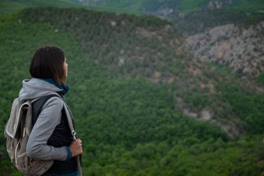 Sırt çantasıyla gezen genç bir kadın ve rahatlatıcı bir ruh haliyle dağın tepesinde oturuyor ve ormanın, mavi gökyüzünün ve tatildeki bulutların güzel manzarasını seyrediyor. Asyalı kadın yalnız seyahat eder..