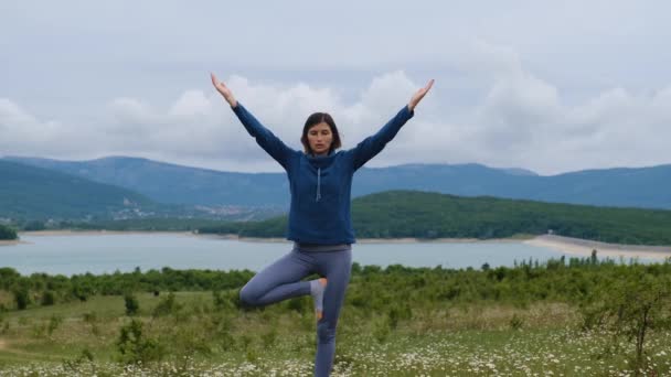 年轻女人在户外放松 她做树的姿势 女人用一条腿支住了嘴 与自然和谐相处 一个女人在湖边的菊花田里做瑜伽 瑜伽运动与健康生活方式概念 — 图库视频影像