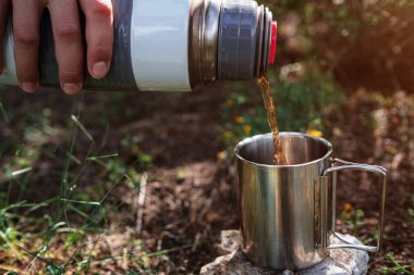 Kamp bardağında dişi bir el, doğadaki bir termostan çay doldurur. Mola, dinlenme ve seyahat fikri