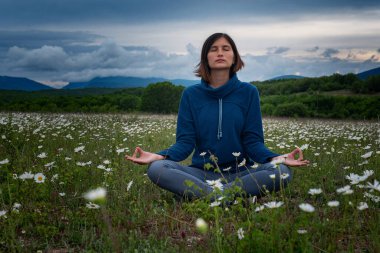 Sahada yoga yapan genç bir kadın. Gün batımından önce bir papatya tarlasında meditasyon. Koyu mavi renkler, sakin ve huzurlu. Ruhani ve duygusal kavram.