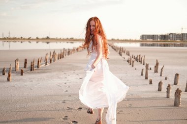 Uzun, sağlıklı, kızıl saçlı ve göl kenarında yürüyen beyaz elbiseli güzel bir bayan. Sıcak yaz akşamı. Gün batımı. Yaşam tarzı kavramı.