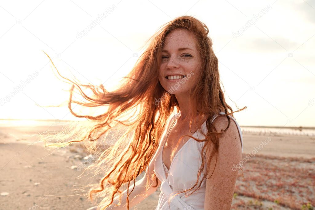Beautiful young lady with long healthy red hair and cute white dress walking near lake. Hot summer evening. Sunset. Lifestyle concept.
