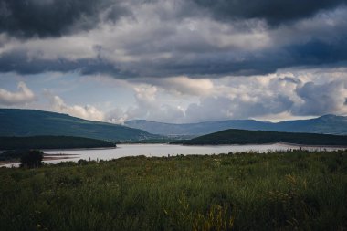 Yağmur yağmadan önce dağların arasında, vadide bir göl. Güzel gün batımı ışığı. Dağ vadisi bulutlu gökyüzü manzarası. Yeşil çalı.