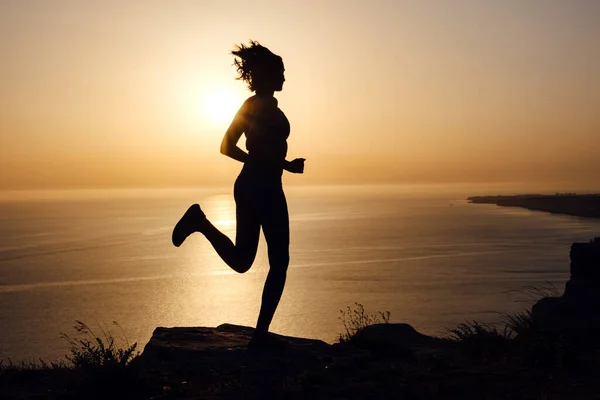 Side view of fitness woman running on a road by the sea. Sportswoman training on seaside promenade at sunset. Summer sport and freedom concept. Athlete training on dusk.