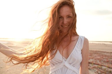 Uzun, sağlıklı, kızıl saçlı ve göl kenarında yürüyen beyaz elbiseli güzel bir bayan. Sıcak yaz akşamı. Gün batımı. Yaşam tarzı kavramı.