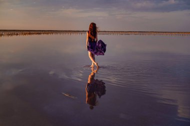 Tuz gölünde yürüyen ipeksi mor elbiseli zarif bir kadın. Romantik bir ruh hali. Bulutların ve boş uzayın su yansıması. Tatil, tatil gezisi sahnesi. Gün batımında görkemli gökyüzü