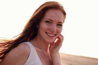 Uzun, sağlıklı, kızıl saçlı ve göl kenarında yürüyen beyaz elbiseli güzel bir bayan. Sıcak yaz akşamı. Gün batımı. Yaşam tarzı kavramı.