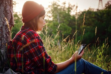 Güzel bayan gezgin ağacın yanında oturuyor ve cep telefonunda daktilo yazıyor. Sonbaharda parkta son moda genç kadın mesajlaşması.