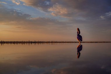 Tuz gölünde yürüyen ipeksi mor elbiseli zarif bir kadın. Romantik bir ruh hali. Bulutların ve boş uzayın su yansıması. Tatil, tatil gezisi sahnesi. Gün batımında görkemli gökyüzü