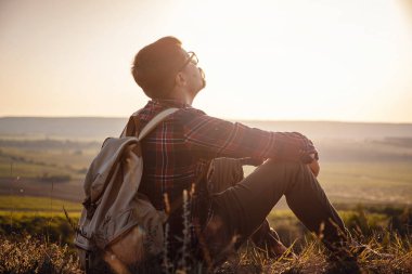 Sırt çantalı bir yürüyüşçü dağın tepesinde oturuyor. Ufukta turuncu gün doğuyor. Yaşam tarzı yürüyüş konsepti, yaz tatilleri.