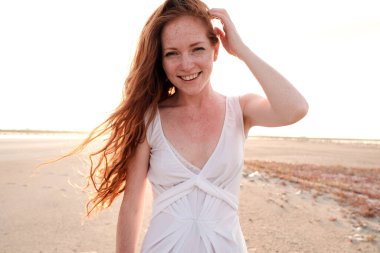 Uzun, sağlıklı, kızıl saçlı ve göl kenarında yürüyen beyaz elbiseli güzel bir bayan. Sıcak yaz akşamı. Gün batımı. Yaşam tarzı kavramı.