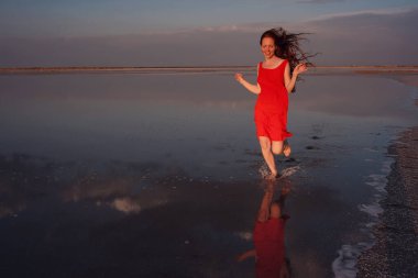 Tuz gölünde yürüyen kırmızı elbiseli zarif bir kadın. Romantik bir ruh hali. Bulutların ve boş uzayın su yansıması. Tatil, tatil gezisi sahnesi. Sivash Gölü