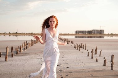 Uzun, sağlıklı, kızıl saçlı ve göl kenarında yürüyen beyaz elbiseli güzel bir bayan. Sıcak yaz akşamı. Gün batımı. Yaşam tarzı kavramı.