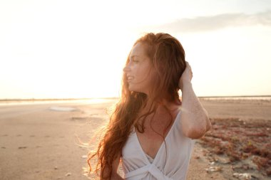 Uzun, sağlıklı, kızıl saçlı ve göl kenarında yürüyen beyaz elbiseli güzel bir bayan. Sıcak yaz akşamı. Gün batımı. Yaşam tarzı kavramı.