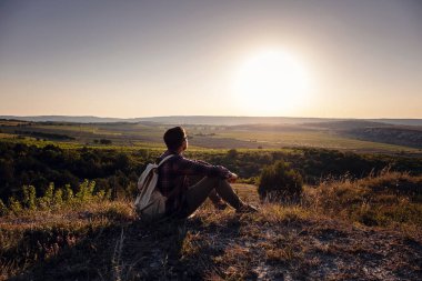 Sırt çantalı bir yürüyüşçü dağın tepesinde oturuyor. Ufukta turuncu gün doğuyor. Yaşam tarzı yürüyüş konsepti, yaz tatilleri.
