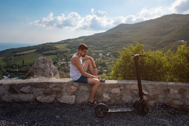 Genç adam dağ sıralarında elektrikli scooter kullanıyor. Uzun bir yolculuktan sonra dinlenmek, temiz havanın ve güzel manzaranın tadını çıkarmak. Ekolojik ulaşım kavramı
