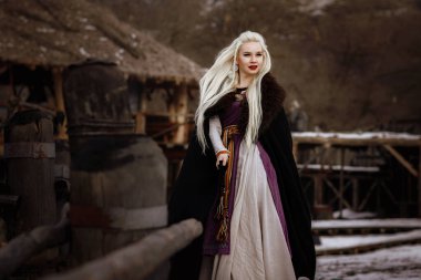 Kalenin zeminine karşı siyah pelerin giymiş güzel sarışın Viking. Güzel bir film seti. Eski bir Viking köyü.