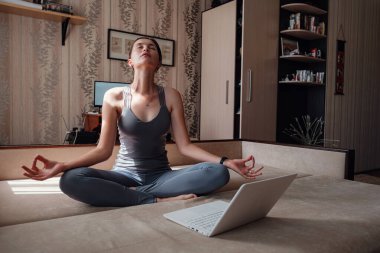 Time for yoga. Attractive young woman exercising and sitting in yoga lotus position while resting at home. View of a woman conducting virtual fitness class at home on a video conference. clipart