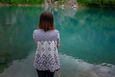 Dağların arasında donduğunu hisseden maceracı Asyalı kadın, doğa manzarasının tadını çıkar. Orman ve göl, panço, boho ve seyahat tutkusu tarzı. Turier Gölü, Alibek Buzulu, Kuzey Kafkasya, Dombai, Rusya.