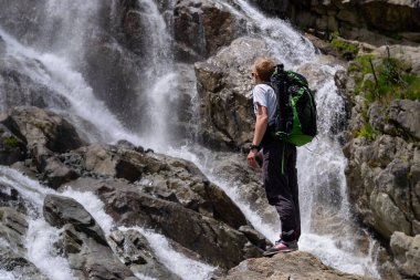 Sırt çantalı bir adam şelaleye bakıyor. Seyahat yaşam tarzı macera konsepti, doğayla uyum içinde aktif tatiller. Kuzey Kafkasya, Dombai, Rusya.