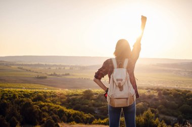Mutlu kadın, dağın eteğinde günbatımının arka planında ellerini kaldırarak duruyor. Genç turist kadın dağlardaki manzaranın tadını çıkarıyor..