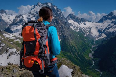 Mavi bluzlu, turuncu sırt çantalı genç bir kadın turist, dağların nefes kesici manzarasının keyfini çıkarıyor. Kuzey Kafkasya, Dombai, Rusya. dağcılık sporları yaşam tarzı konsepti