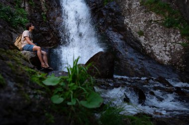 Sırt çantalı yürüyüşçü waalkinh güzel doğa manzarasında parktaki şelaleye bakıyor. Açık havada duran yetişkin erkek portresi. Şelale Shumka, Dombai, Rusya