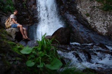Sırt çantalı yürüyüşçü waalkinh güzel doğa manzarasında parktaki şelaleye bakıyor. Açık havada duran yetişkin erkek portresi. Şelale Shumka, Dombai, Rusya