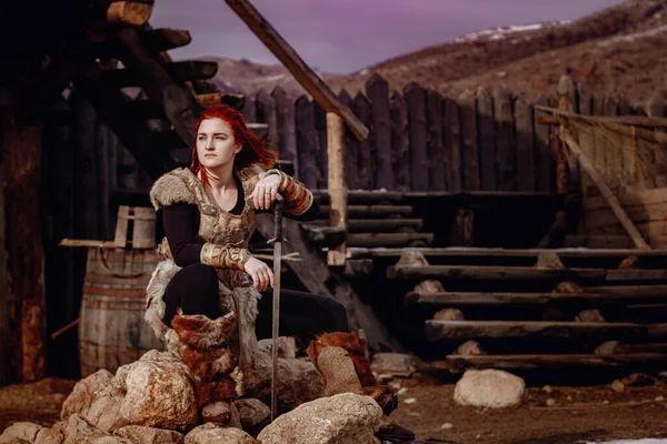 赤い髪のバイキングの女の子 剣の手に敵と戦うために 中世の風景の再構築 大規模なバイキング村を背景に — ストック写真