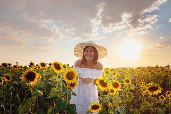 Beautiful young girl enjoying nature on the field of sunflowers at sunset. Asian girl in a cute white dress and hat enjoys summer and vacation.